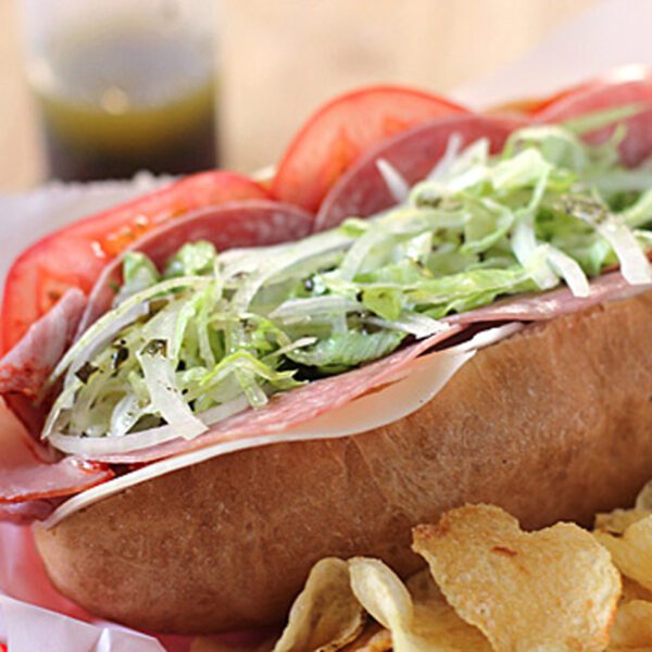 A submarine sandwich filled with sliced deli meats, cheese, shredded lettuce, and tomato sits in a red basket next to potato chips. A small bottle of dressing is visible in the background.