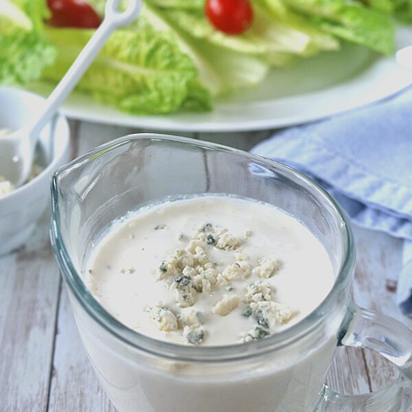 A glass measuring cup filled with creamy blue cheese dressing sits on a wooden table, with crumbled blue cheese on top. In the background, a plate of fresh romaine lettuce and cherry tomatoes is visible.