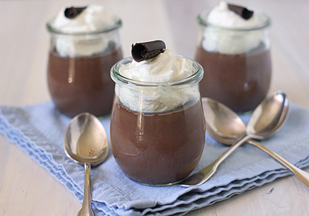 Three glass jars filled with chocolate mousse, topped with whipped cream and a chocolate curl, are placed on a folded blue cloth, surrounded by three metal spoons.