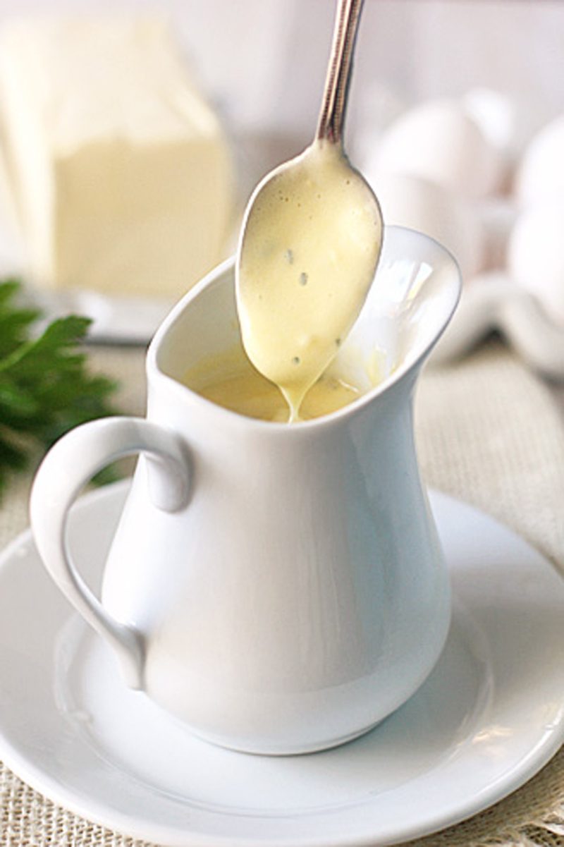 A spoon drizzles creamy yellow sauce, possibly hollandaise, into a white ceramic pitcher on a matching saucer. Butter, eggs, and parsley are blurred in the background.
