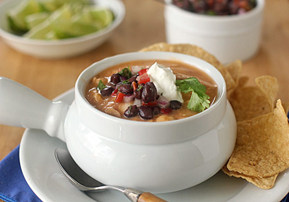 A bowl of creamy soup topped with black beans, diced tomatoes, cilantro, and a dollop of sour cream, served with tortilla chips on the side. A spoon rests on the plate, with lime wedges and salsa in the background.