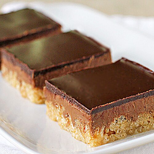 Three chocolate-topped dessert bars with a thick chocolate layer over a crumbly, light-colored base, arranged in a row on a white rectangular plate.