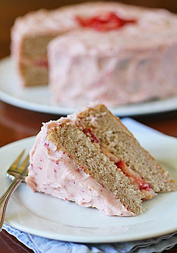A slice of layered cake with pink strawberry frosting sits on a white plate with a fork, with the rest of the cake in the background on a cake stand.