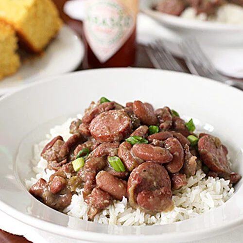 A white bowl filled with white rice, topped with red beans and sausage slices, garnished with green onions. In the background, there is cornbread, a bottle of Tabasco sauce, and a fork and knife.