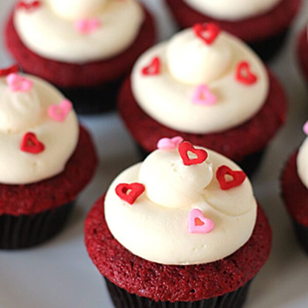 Red velvet cupcakes topped with swirls of white frosting and decorated with red and pink heart-shaped sprinkles, arranged on a white plate.