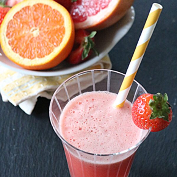 A glass of pink fruit juice with a yellow-and-white striped straw and a strawberry garnish. Behind it, a plate holds a halved grapefruit, an orange slice, and strawberries.