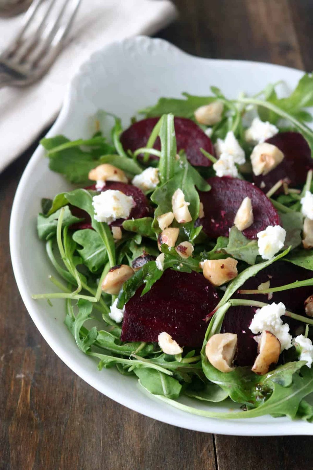 A white bowl filled with arugula, roasted beet slices, crumbled goat cheese, and chopped nuts sits on a dark wooden table with a napkin and two forks in the background.