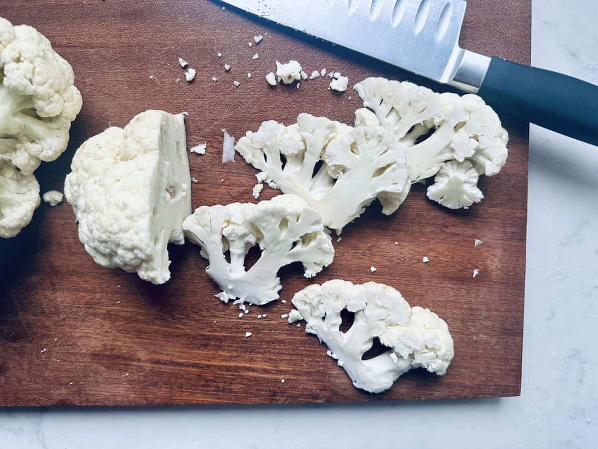 Several slices of raw cauliflower and a knife rest on a dark wooden cutting board, with small cauliflower pieces scattered around. Part of a whole cauliflower is visible on the left side of the board.