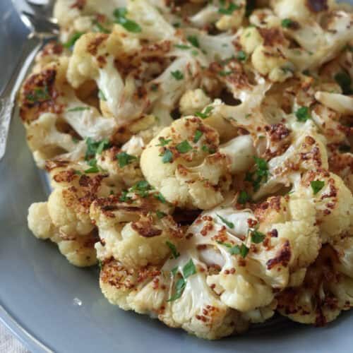 A plate of roasted cauliflower florets garnished with chopped herbs, served on a gray dish with a silver serving spoon on the side.