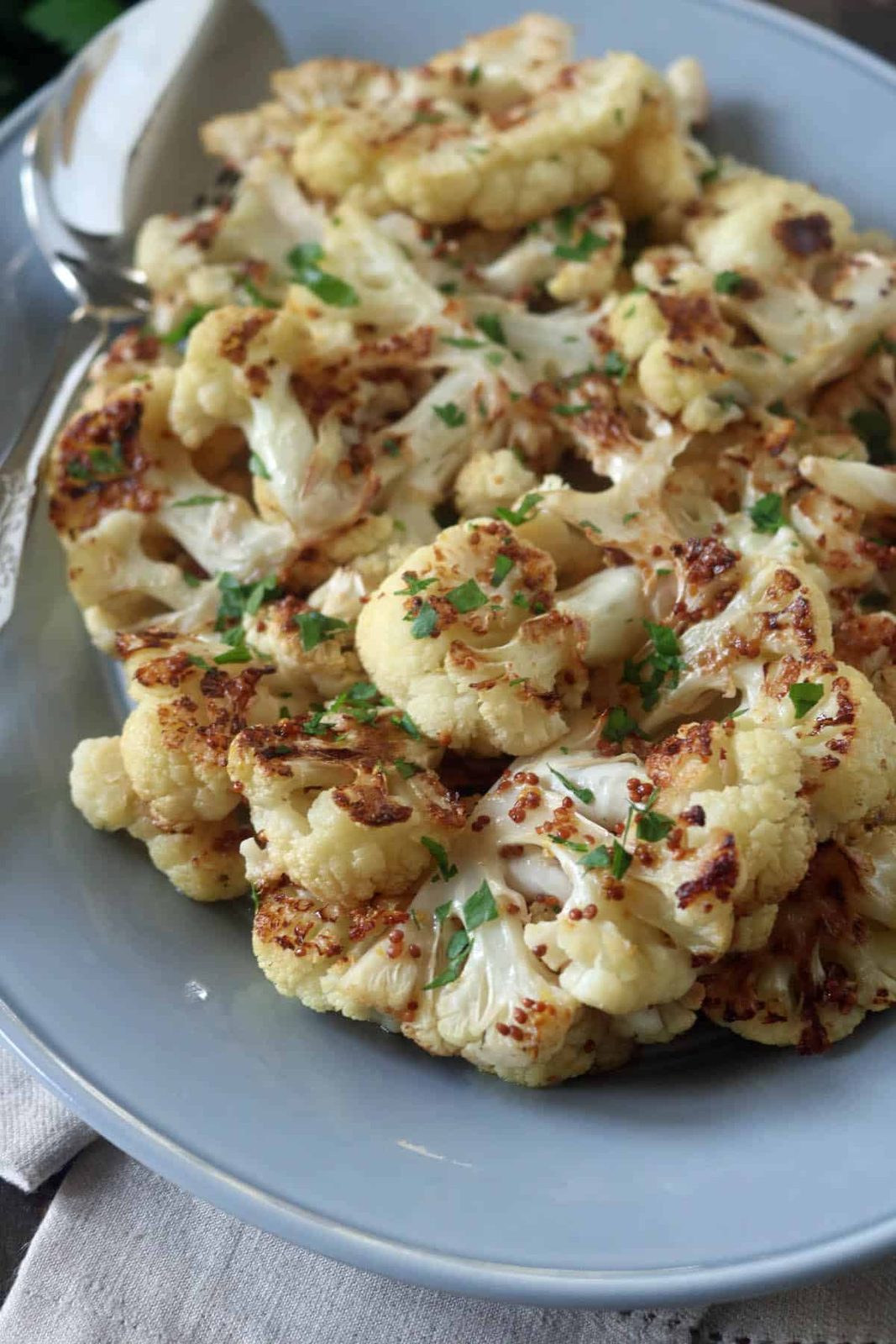 A plate of roasted cauliflower florets garnished with chopped herbs, served on a gray dish with a silver serving spoon on the side.