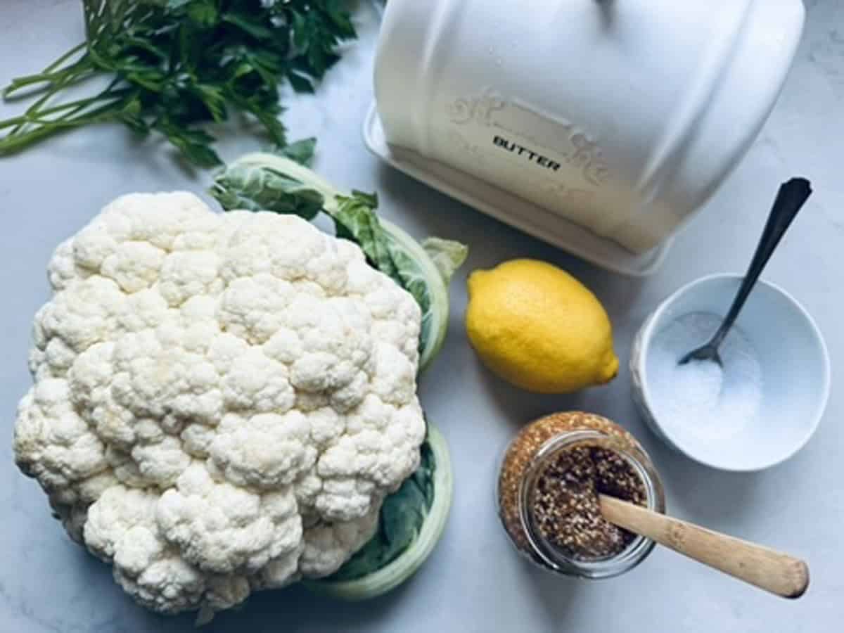 A head of cauliflower, a bunch of parsley, a lemon, a butter dish, a bowl of salt with a spoon, and a jar of grainy mustard are arranged on a white countertop.