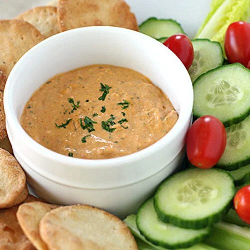 A bowl of creamy dip garnished with herbs is surrounded by pita chips, cucumber slices, cherry tomatoes, celery sticks, and lettuce on a white plate.
