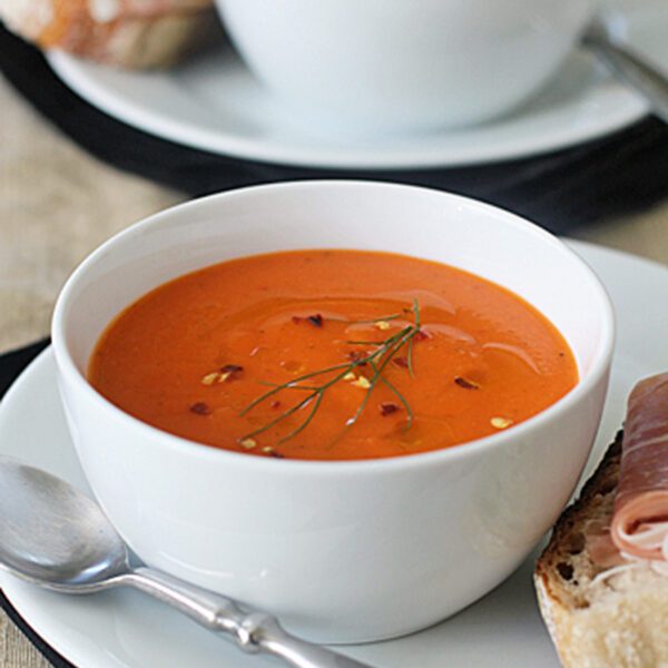 A white bowl of creamy tomato soup garnished with herbs and chili flakes sits on a white plate with a spoon. Next to it is bread topped with prosciutto; another similar setting is blurred in the background.