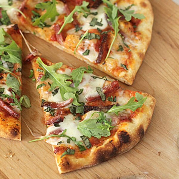A sliced pizza on a wooden board topped with melted cheese, tomato sauce, fresh basil, and arugula leaves. One slice is slightly pulled out from the rest.
