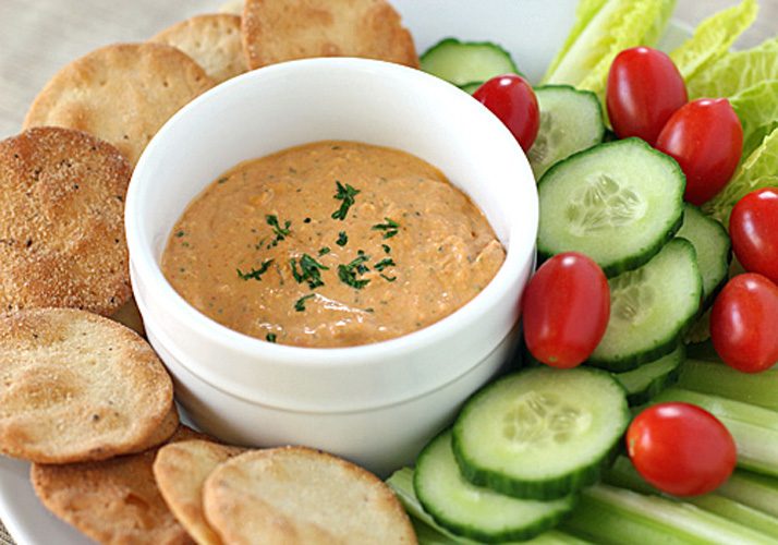 A bowl of creamy dip garnished with herbs is surrounded by pita chips, cucumber slices, cherry tomatoes, celery sticks, and lettuce on a white plate.