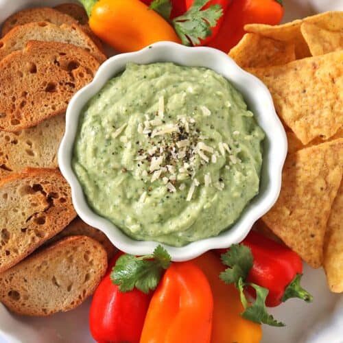 A bowl of green dip topped with shredded cheese and pepper, surrounded by sliced bread, tortilla chips, mini bell peppers, and cilantro on a white plate.