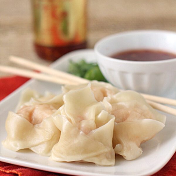 A plate of steamed dumplings with chopsticks rests on an orange napkin, accompanied by a bowl of dipping sauce and a jar of chili sauce in the background.