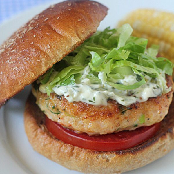 A chicken burger with lettuce, tomato, and creamy sauce on a toasted bun, served on a white plate with a side of corn on the cob.