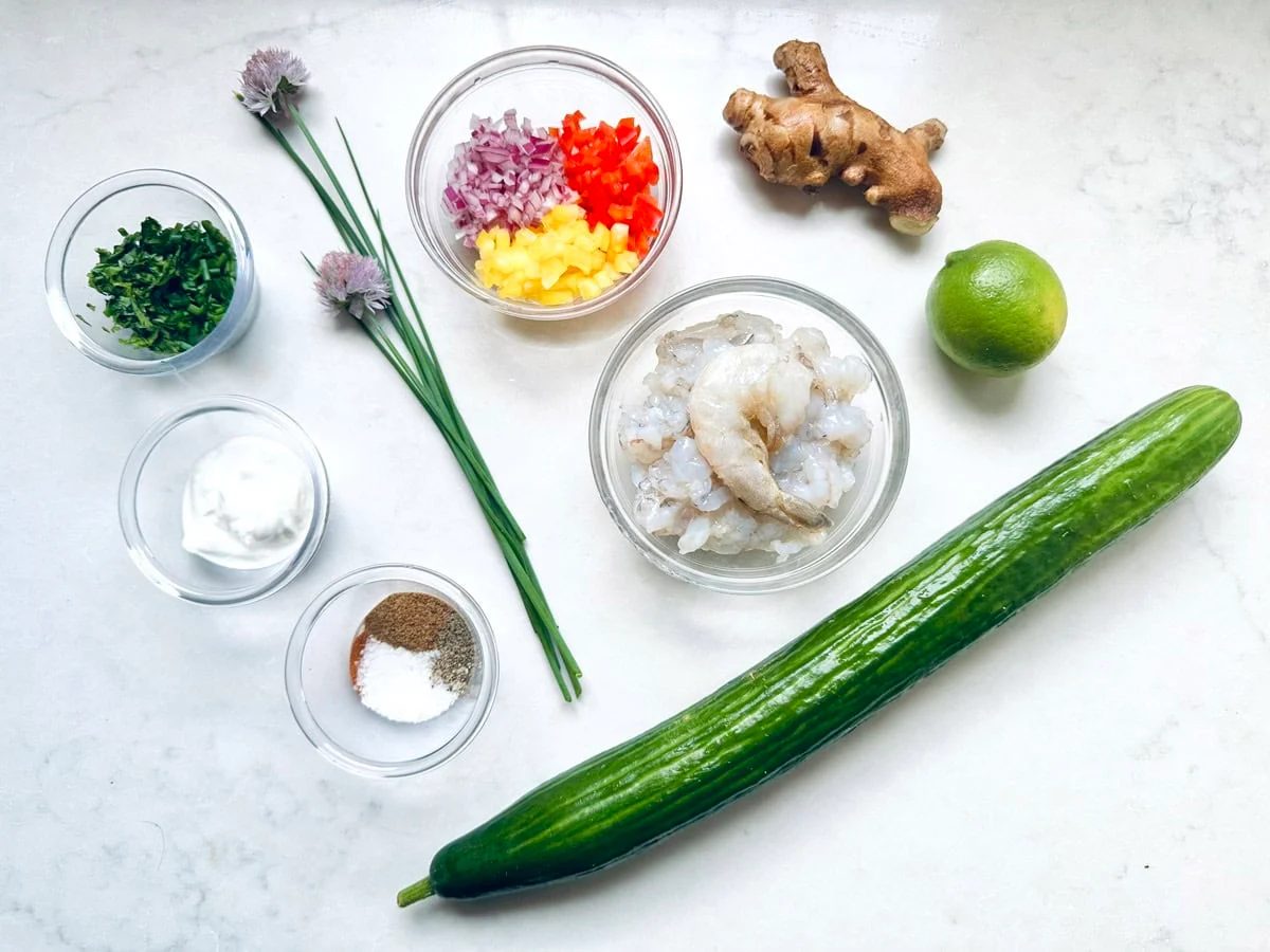 Top-down view of cooking ingredients on a white surface, including cucumber, lime, ginger, shrimp, chopped herbs, chopped red onion and bell pepper, yogurt, and small bowls of salt and pepper.