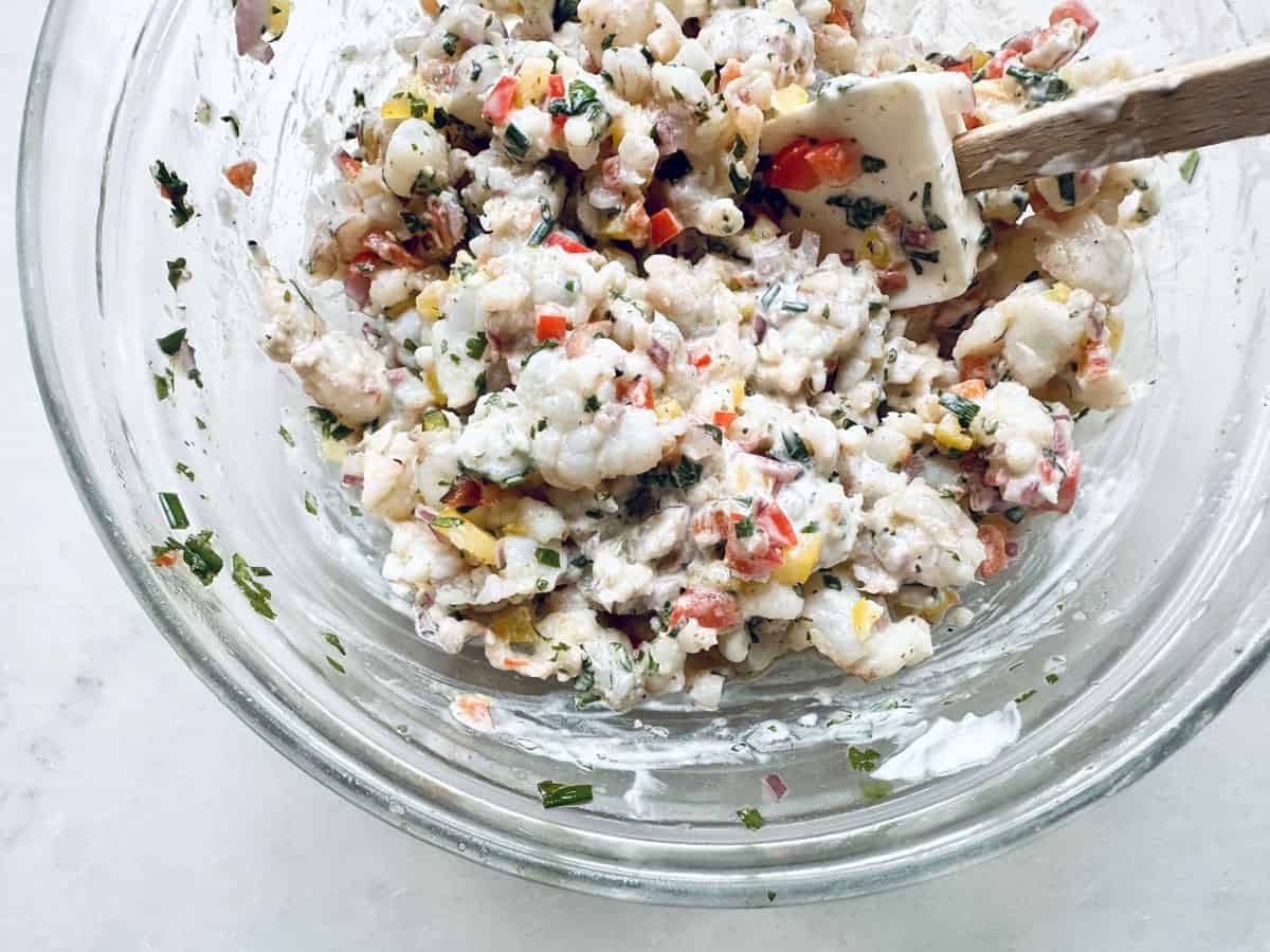 A glass bowl filled with a creamy shrimp salad mixed with chopped red and yellow bell peppers and fresh herbs, being stirred with a white spatula.