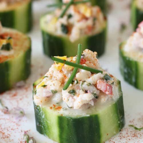 Close-up of cucumber slices topped with a creamy seafood salad mixture, garnished with chives and a sprinkle of paprika, arranged on a white surface.