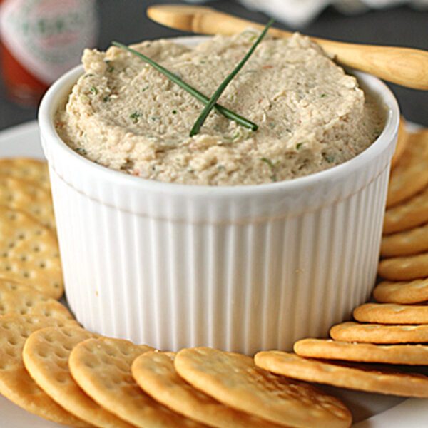 A white ramekin filled with a creamy dip, garnished with two chive stalks, surrounded by round crackers on a white plate. A wooden spreader and a bottle of hot sauce are visible in the background.
