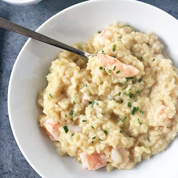A bowl of creamy seafood risotto garnished with chopped chives sits on a gray surface. A fork rests in the risotto, and a small bowl of lemon wedges is placed nearby.