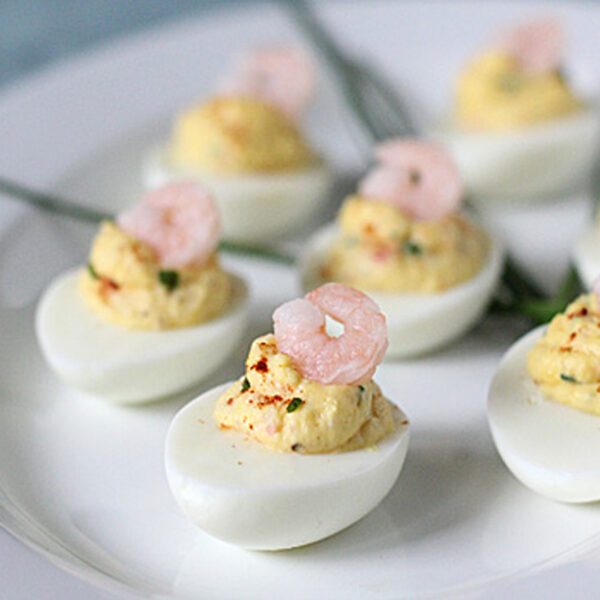 A white plate with several deviled eggs, each topped with a small shrimp and garnished with herbs. The eggs are neatly arranged, and there are some green chives in the background.