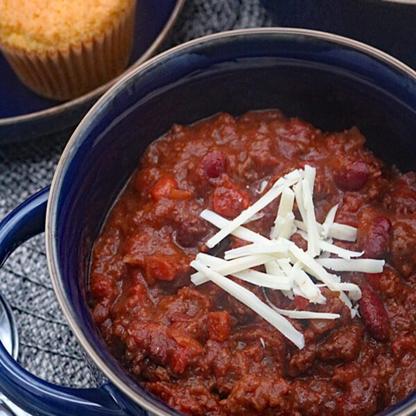 A bowl of chili topped with shredded cheese sits next to a plate of cornbread muffins, with another bowl of chili partially visible in the background.