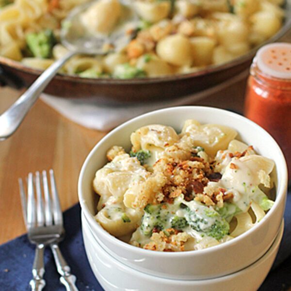 A bowl of pasta shells with broccoli in a creamy sauce, topped with crispy breadcrumbs, sits on a blue napkin with two forks. A skillet with more pasta and a shaker of red spice are in the background.