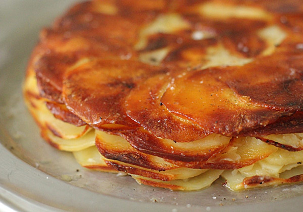 A close-up of a golden-brown potato galette, showing thin, crispy layers of sliced potatoes baked together and neatly arranged in a circular pattern on a metal plate.