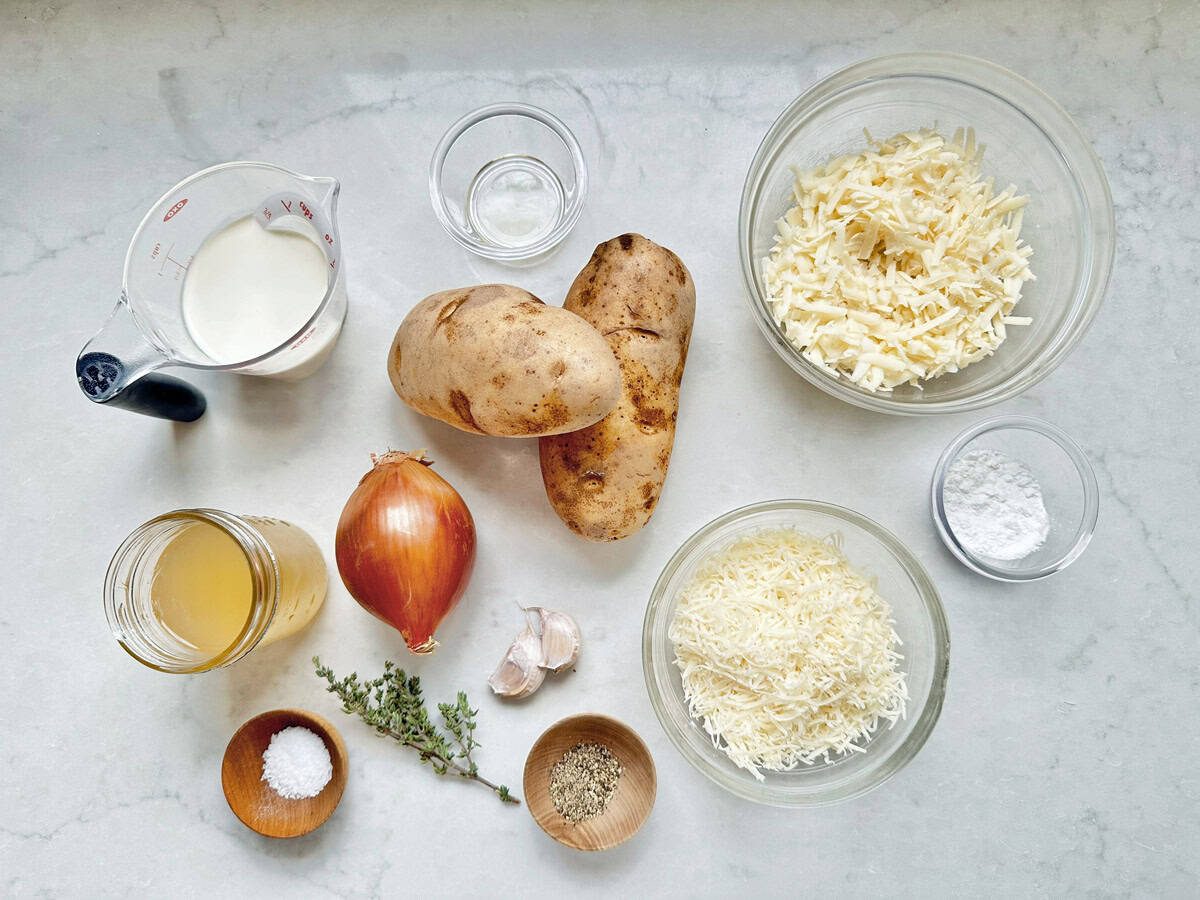 Overhead view of ingredients on a white surface: two russet potatoes, shredded cheese in two bowls, milk in a measuring cup, an onion, two garlic cloves, fresh thyme, chicken broth in a jar, flour, salt, and pepper in small bowls.