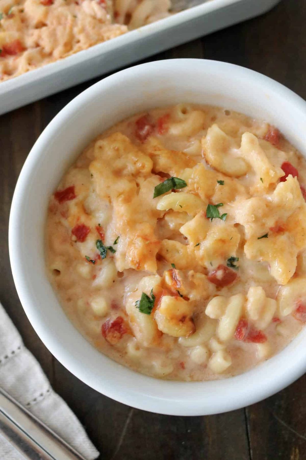 A white bowl filled with creamy macaroni and cheese mixed with diced tomatoes and topped with melted cheese and chopped herbs, sitting on a wooden table.