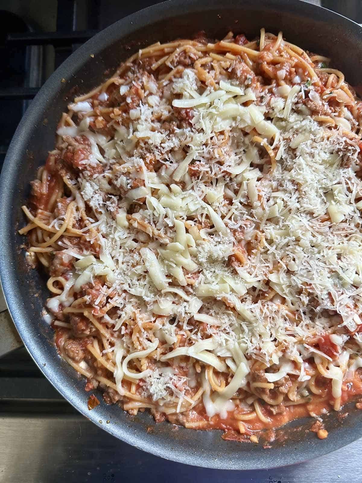 A close-up of a skillet filled with spaghetti in a tomato meat sauce, topped with shredded mozzarella and grated parmesan cheese, cooking on a stovetop.