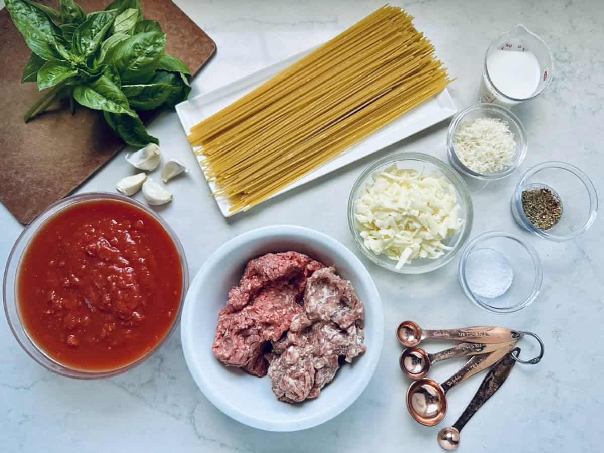 A top-down view of ingredients for a pasta dish: uncooked spaghetti, ground meat, tomato sauce, grated cheese, milk, basil leaves, garlic, spices, salt, and measuring spoons arranged on a white counter.