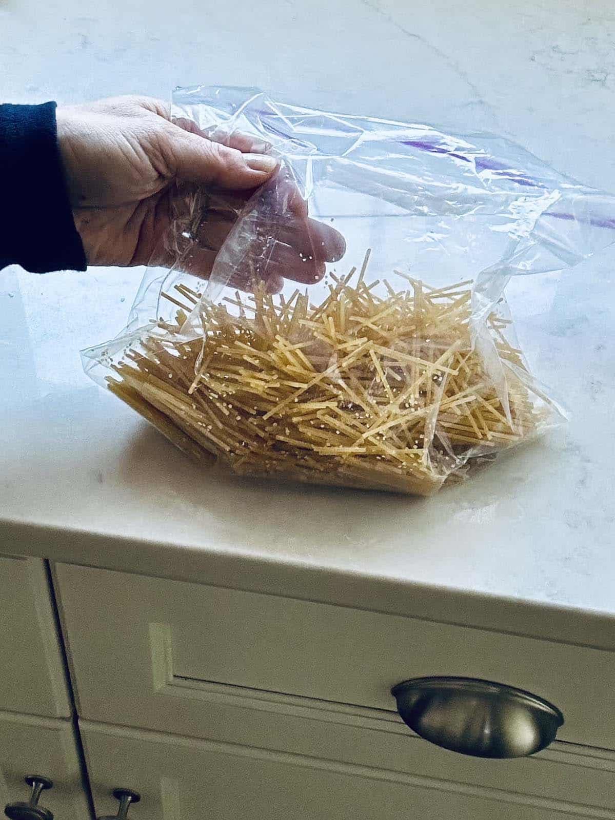 A hand holds a clear plastic bag filled with broken pieces of uncooked spaghetti noodles over a white kitchen countertop.