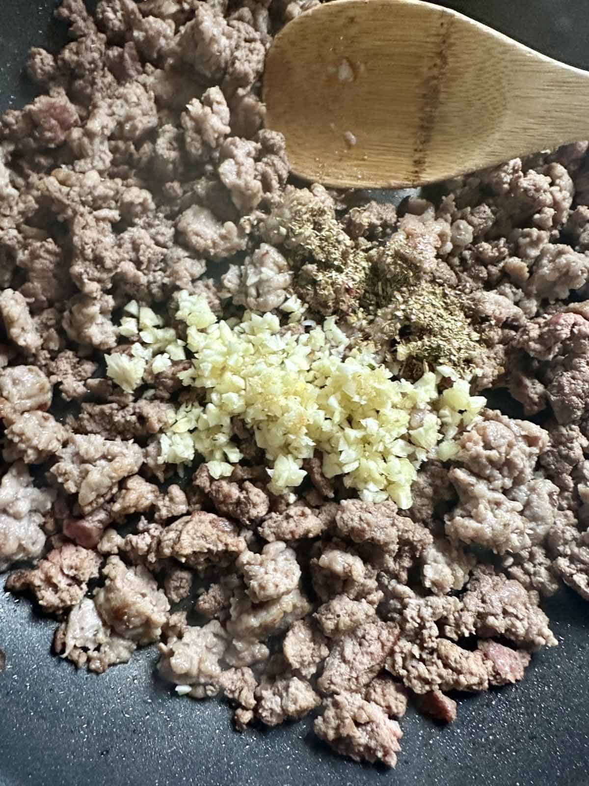 Ground beef cooking in a pan, topped with chopped garlic and dried herbs, being stirred with a wooden spoon.