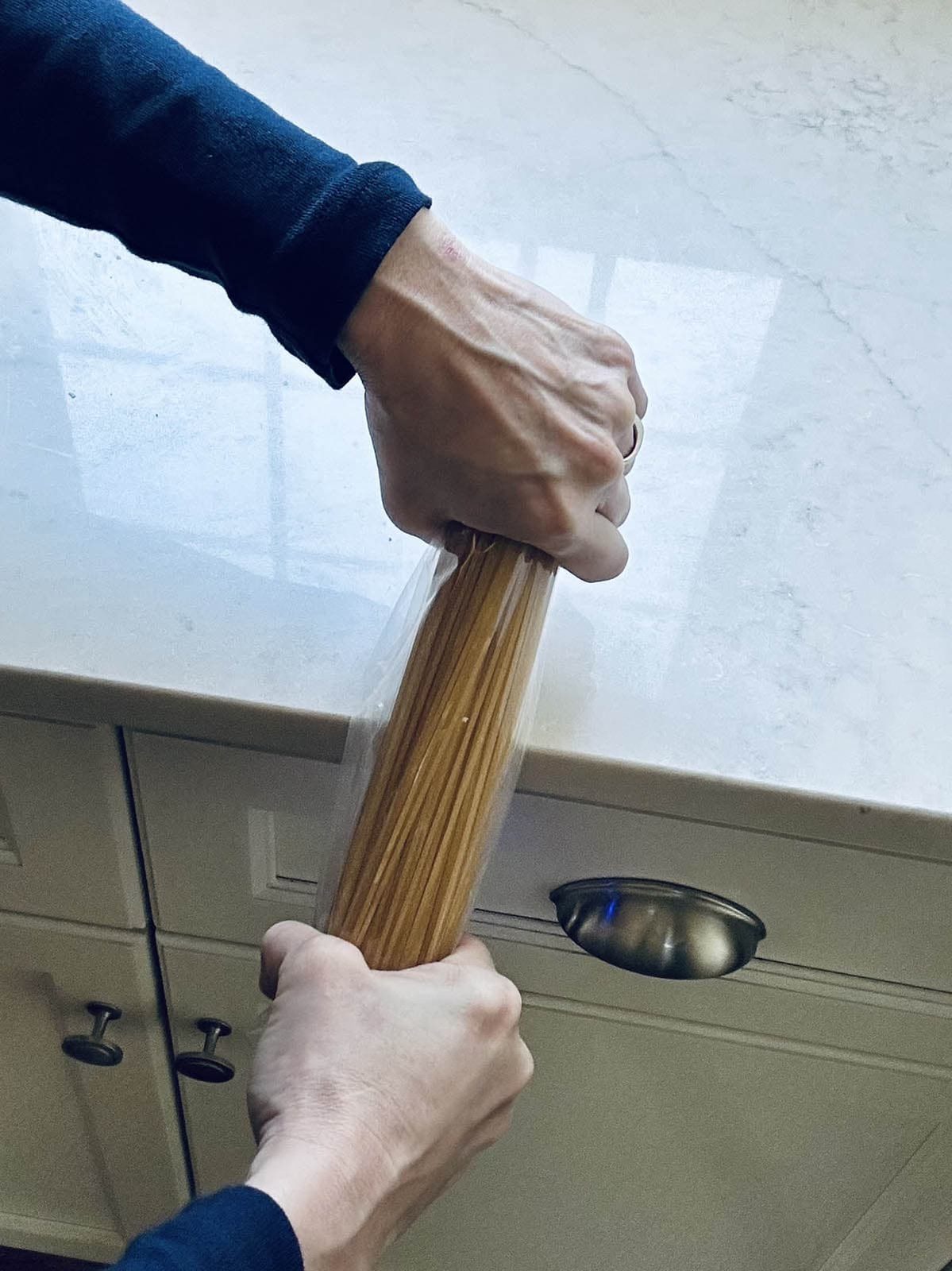 Two hands are seen breaking a bundle of uncooked spaghetti in half over a kitchen counter with white cabinets below.