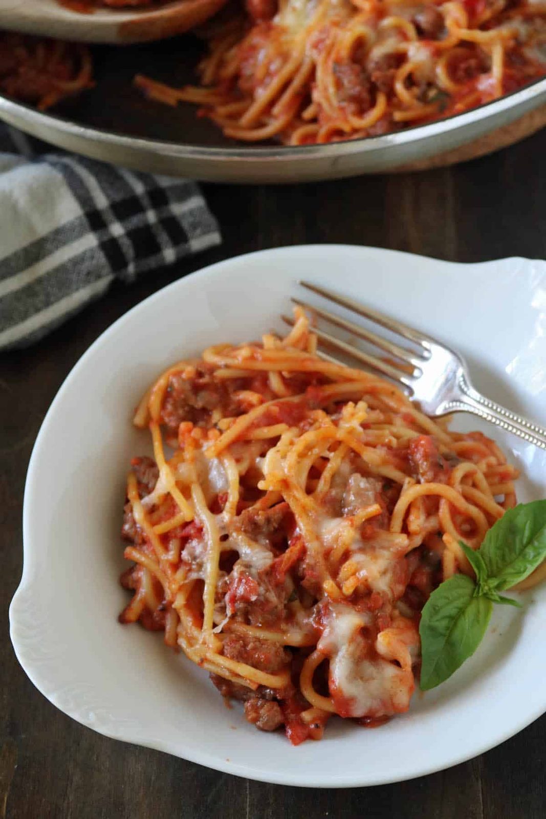 A white plate with baked spaghetti topped with melted cheese, ground meat, and tomato sauce. A fork rests on the plate, and a fresh basil leaf is placed as garnish. Part of a pan and a cloth napkin appear in the background.