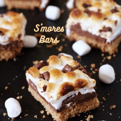 Close-up of S’mores bars with toasted marshmallows, melted chocolate, and a graham cracker crust, arranged on a dark surface with marshmallows scattered around.