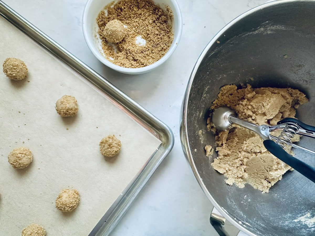A baking tray lined with parchment paper holds cookie dough balls. Nearby, a bowl with cookie dough and a scoop, and a small bowl of crumbs are visible on a white surface.