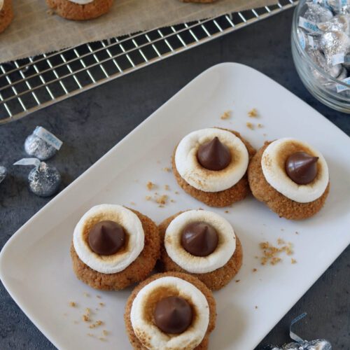Five cookies topped with marshmallow rings and chocolate kisses are arranged on a white plate, with more cookies on a cooling rack and wrapped chocolate kisses nearby on a gray surface.