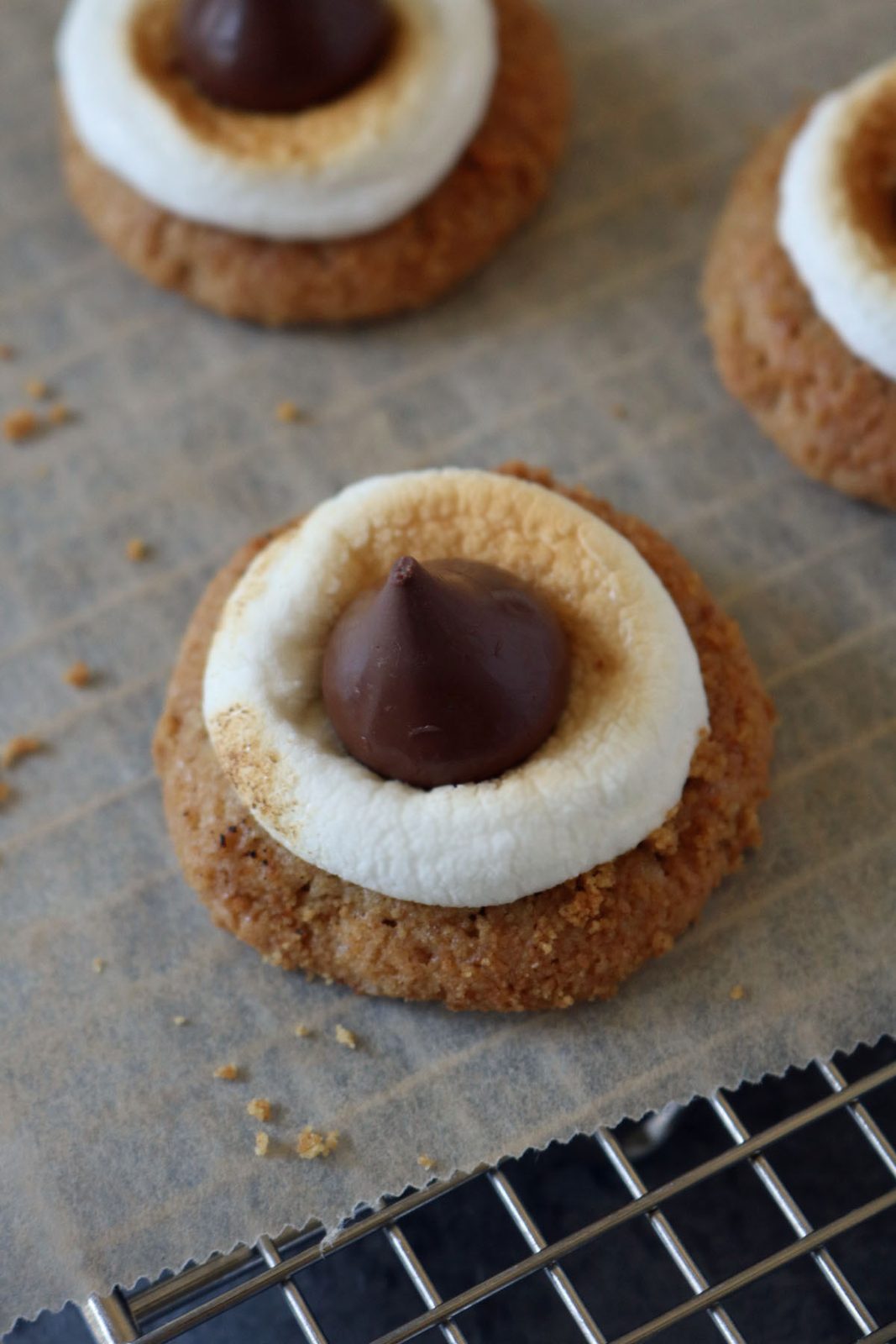 A close-up of a cookie topped with a toasted marshmallow ring and a chocolate Hersheys Kiss, resting on parchment paper above a wire cooling rack.