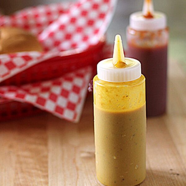 A squeeze bottle of yellow mustard sits on a wooden surface, with a bottle of red sauce and a red-checkered food basket blurred in the background.