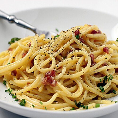 A plate of spaghetti carbonara topped with finely chopped parsley, crispy bacon bits, and a sprinkle of black pepper, with a fork resting on the side of the white bowl.