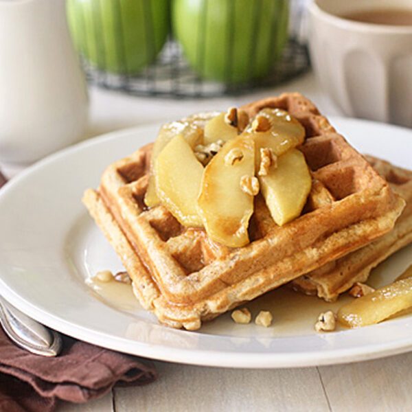 A plate with two stacked waffles topped with cooked apple slices and chopped nuts. A fork and napkin are beside the plate, with a cup of coffee and a small pitcher in the background. Green apples are also visible.