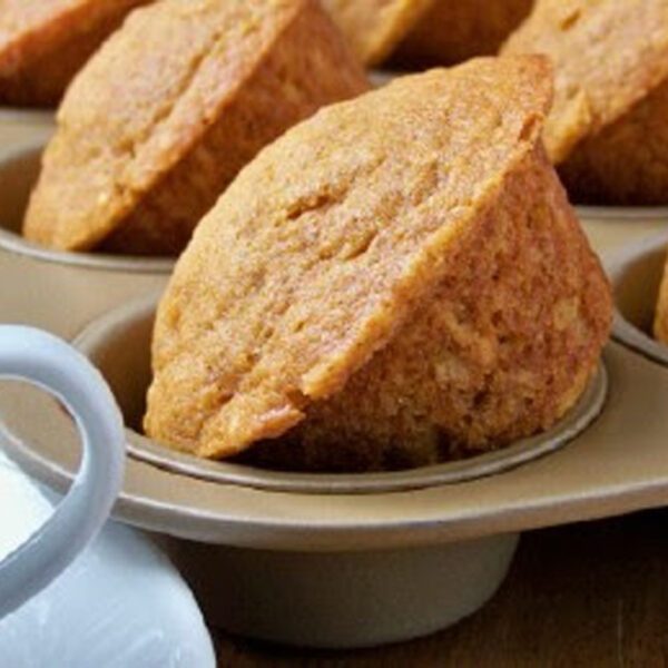 A close-up of freshly baked muffins in a muffin tin, with a white coffee cup partially visible in the foreground. The muffins are golden brown and slightly domed on top.