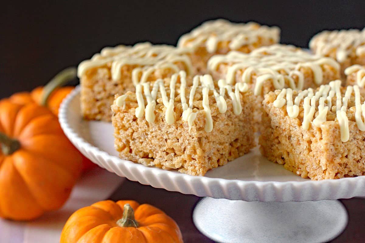 Fluffer-nutter Treats: Rice Krispies treats squares drizzled with white icing are displayed on a white cake stand, surrounded by small orange pumpkins.