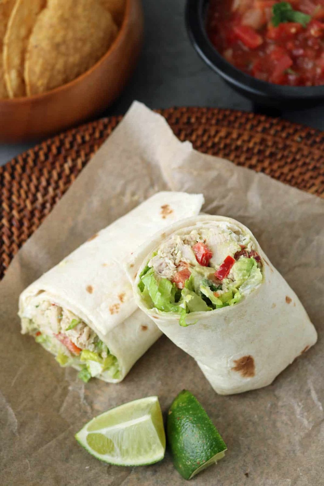 A Spicy Chicken Salad with Chipotle and Avocado wrap, cut in half and filled with lettuce and red bell peppers, sits on parchment paper with lime wedges. Tortilla chips in a wooden bowl and a bowl of salsa are in the background.
