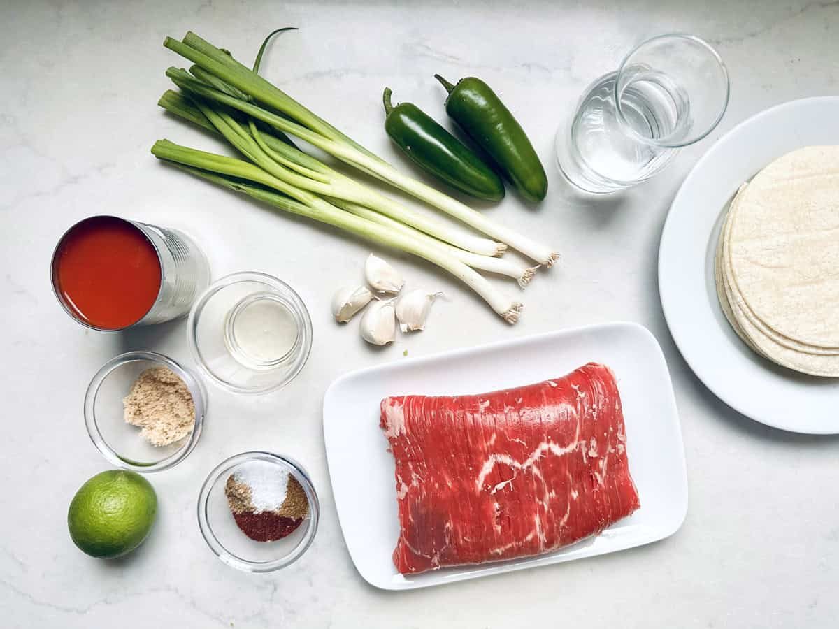 Top-down view of ingredients on a white surface: green onions, two jalapeños, a glass of water, corn tortillas, a plate of raw steak, garlic cloves, a can of tomato sauce, lime, spices, and bowls of brown sugar and oil.